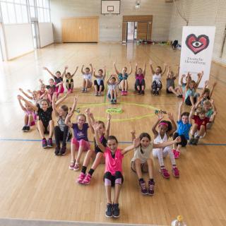 Eine Gruppe Kinder sitzt herzförmig angeordnet in einer Sporthalle