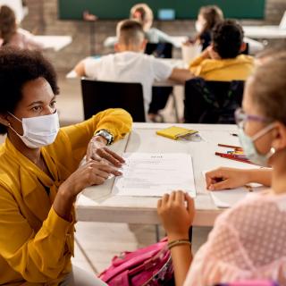 Unterrichtssituation: Lehrerin mit medizinischer Maske erklärt einer Schülerin etwas (die Schülerin trägt ebenfalls eine medizinische Maske). 