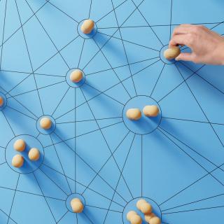 Hölzerne Spielfiguren stehen auf blauem Untergrund, auf dem ein Netzwerk aus mit Linien verbundenen Punkten gezeichnet ist. Eine Hand berührt eine der Spielfiguren.