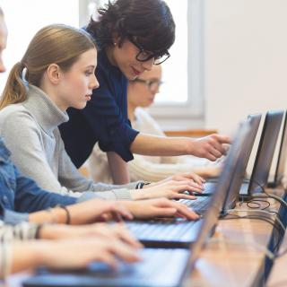 Schülerinnen und Lehrkraft arbeiten jeweils an einem Laptop.