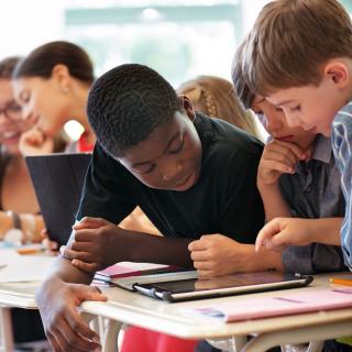 Schülerinnen und Schüler blicken auf Tablets, die vor ihnen auf dem Tisch liegen.