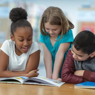 Grundschülerinnen und Grundschüler sitzen an einem Tisch vor aufgeschlagenen Büchern, ein Mädchen liest vor und die anderen hören zu.