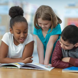 Grundschülerinnen und Grundschüler sitzen an einem Tisch vor aufgeschlagenen Büchern, ein Mädchen liest vor und die anderen hören zu.
