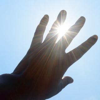Eine linke Hand vor blauem Himmel, durch die Finger blinzelt die Sonne hindurch.