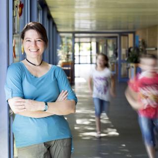 Lehrerin steht lächelnd in einem Schulflur, unscharf  im Hintergrund zu erkennen sind laufende Schülerinnen und Schüler.