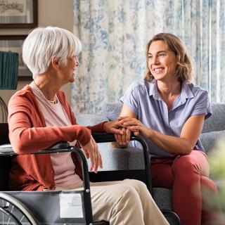 Eine jüngere Frau und eine ältere, in einem Rollstuhl sitzende, ältere Frau halten sich lächelnd an den Händen.