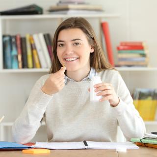 Jugendliche sitzt an einem Schreibtisch, in den Händen eine Tablette und ein Trinkgefäß.