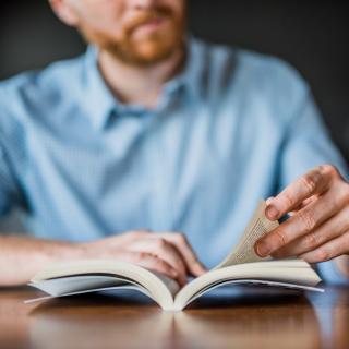 Nahaufnahme: Auf einem Tisch liegt ein geöfffnetes Buch, eine Hand hält es offen, die andere blättert darin. Im Hintergrund ist unscharf die lesende Person zu erkennen.