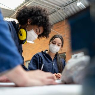 Jugendliche in Arbeitskleidung arbeiten gemeinsam über einen Tisch gebeugt. 