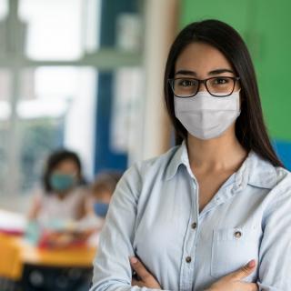 Lehrerin in einem Klassenraum, sie trägt einen medizinischen Mund-Nasen-Schutz.