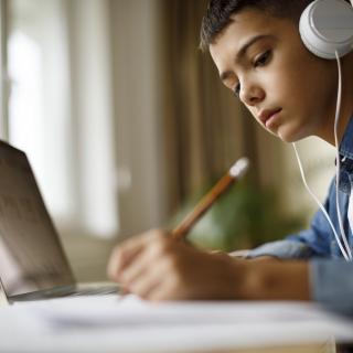 Ein junge sitzt schreibend und Kopfhörer tragend an einem Schreibtisch vor einem geöffneten Laptop.