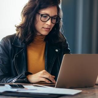 Eine Frau sitzt an einem Schreibtisch und schaut in einen aufgeklappten Laptop.