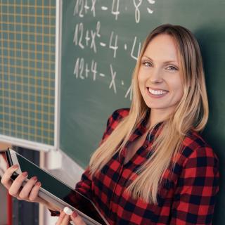 Eine Lehrerin steht mit dem Rücken zur Tafel, in ihren Händen hält sie einen Tablet-Computer.