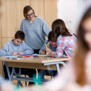 Unterrichtssituation in einer Grundschule: Schülerinnen und Schüler arbeiten, eine Lehrerin blickt ihnen über die Schulter. 