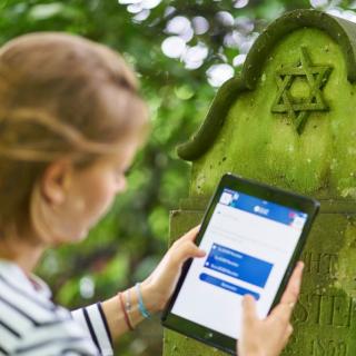 Eine Schülerin mit Tablet steht vor einem jüdischen Grabstein.