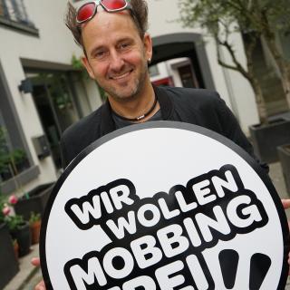 Der Comedian und Musiker Tom Lehel hält das Logo der Initiative "Wir wollen mobbingfrei!" in den Händen.