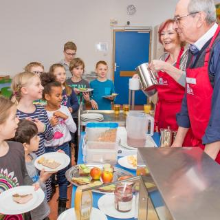 Kinder stehen vor einem Tresen, an dem zwei Erwachsene Frühstück ausgeben.