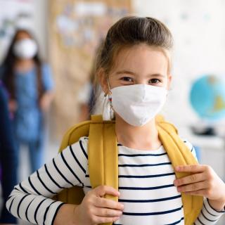 Eine Schülerin mit Maske und Tornister steht in der Schule