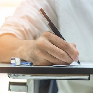 Detailaufnahme einer Hand, die auf Papier schreibt. Im Hintergrund unscharf weitere schreibende Personen an Tischen.