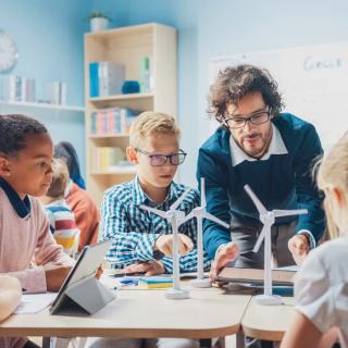 Lehrer mit Schülerinnen und Schülern in einer Unterrichtssituation. Auf dem Tisch stehen Modellwindräder, die die Kinder und der Lehrer gemeinsam betrachten.