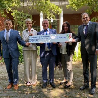 Herr Eichhorst, Ministerin Feller, Herr Schulte, Frau Guhl und Herr Geldmacher stehen mit der LAQUILA-Absenderkennung auf einem Schild in einem begrünten Innenhof