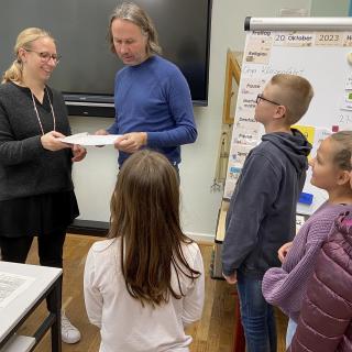 In einem Klassenraum überreicht Lehrerin Anna Müller dem Alltagshelfer Christian Böhle Papiere. Im Vordergrund stehen Schülerinnen und Schüler. 