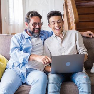 Vater und Sohn sitzen auf einem Sofa und schauen lächelnd in einen geöffneten Laptop auf dem Schoß des Sohnes.