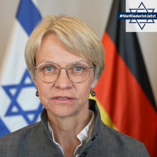 Portrait von Schul- und Bildungsministerin Dorothee Feller, im rechten oberen Bildrand ist eine Grafik des Davidsterns mit dem Schriftzug #NieWiederIstJetzt eingefügt, im Hintergrund sind die Flaggen Europas, Israels und Deutschlands erkennbar.