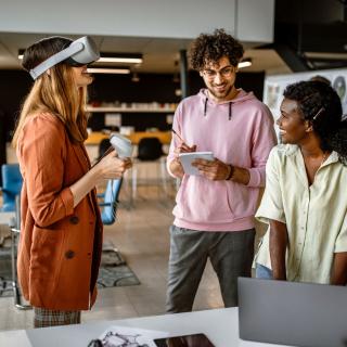 Drei junge Menschen arbeiten mit verschiedenen Medien, unter anderem mit einer VR-Brille.