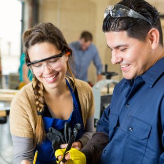Eine Schülerin und ein Lehrer stehen zusammen in einer Werkstatt, sie tragen Arbeitskleidung und Schutzbrillen.
