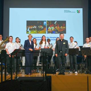 Hauptschultag in Essen: Ministerin Dorothee Feller steht mit Schülerinnen und Schülern auf einer Bühne. Die Jugendlichen halten Musikinstrumente in den Händen.