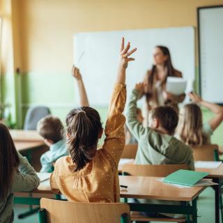 Rückansicht von Grundschülerinnen und Grundschülern, die in einem Klassenraum an ihren Tischen sitzen. Im Hintergrund ist unscharf eine Lehrerin zu erkennen.
