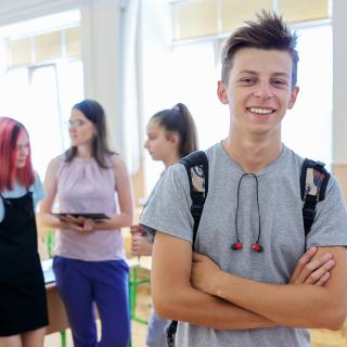 Eine Gruppe Schülerinnen und Schüler steht in einem Klassenraum und spricht miteinander. Ein Jugendlicher im Vordergrund blickt lächelnd in die Kamera.