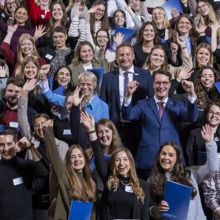 Gruppenbild: Ministerpräsident Hendrik Wüst und Ministerin Dorothee Feller umringt von Referendarinnen und Referendaren im Treppenhaus der Bezirksregierung Düsseldorf. Die Referendarinnen und Referendare halten Mappen mit ihren Urkunden in den Händen.