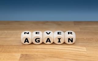 Auf einer Holzplatte liegen fünf Buchstabenwürfel, die die englischen Worte "NEVER" und "AGAIN" bilden.