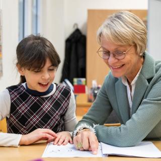 Ministerin Feller und eine Grundschülerin sitzen an einem Tisch und lesen gemeinsam in einem Buch.
