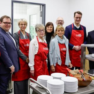 Ministerin Feller, Uschi Glas und Ministerpräsident Wüst stehen mit ehrenamtlichen Mitarbeitenden von "BrotZeit" in einer Schulküche hinter einer Ausgabe mit Frühstückszutaten.