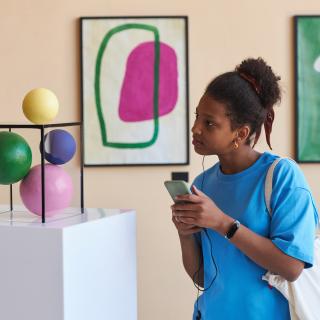 Eine Schülerin steht in einem Museum mit dem Handy in der Hand und einem Kopfhörer im Ohr vor einer Skulptur.