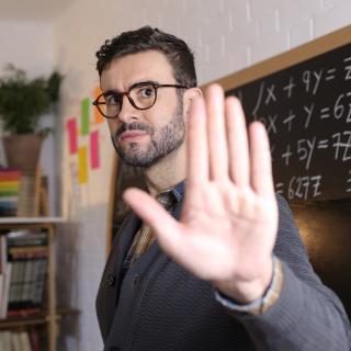 Lehrer steht mit zur Abwehr erhobener Hand vor der Tafel.