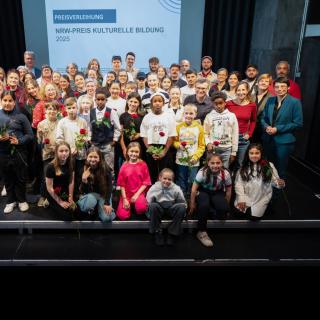 Schülerinnen und Schüler stehen, knien und hocken auf einer Bühne vor einem Banner zur Verleihung des NRW-Preises Kulturelle Bildung 2025