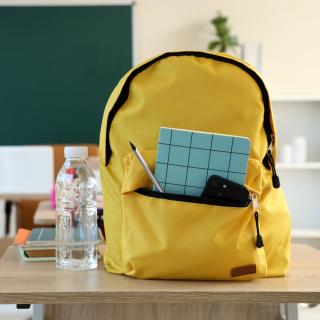 Symbolbild: Rucksack steht auf einem Schultisch, ein Smartphone und Schreibutensilien ragen aus einer geöffneten Tasche.