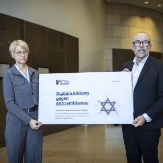 Ministerin Feller und Bert Römgens präsentieren ein Plakat zum Start von AMUDIM (25. Februar 2026)