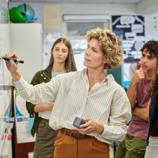 Eine Lehrerin steht an einer Tafel und erklärt den um sie herumstehenden Schülerinnen und Schülern einen Sachverhalt.