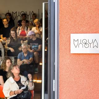 Durch die geöffnete Tür blickt man ins MiQua.forum, in dem vielen Menschen an einer Veranstaltung teilnehmen.