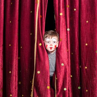 Ein Junge mit aufgemalten Clownsgesicht steckt den Kopf durch einen roten Vorhang.