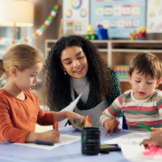 Symbolbild: Lehrerin spricht mit einer Gruppe Kinder, die an einem Tisch sitzen.
