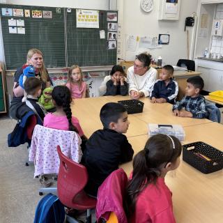 Lehrerin sitzt mit einer Handpuppe vor Kindern, die an einem Tisch sitzen.