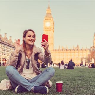 Eine Schülerin sitzt auf einer Wiese und winkt in ihr Smartphone, im Hintergrund sind die Houses of Parliament und Big Ben in London zu erkennen.
