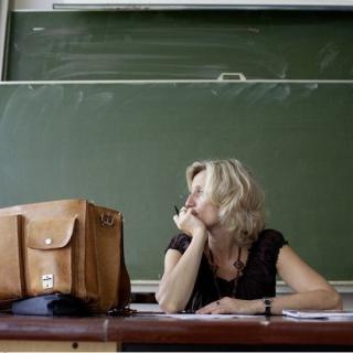 Lehrerin sitzt nachdenklich am Tisch vor einer Tafel.