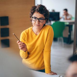 Frau setzt zum Sprechen an, dabei stützt sie sich auf einen Tisch auf. Ihre Gesprächspartner sind nur von Hinten zu erkennen. 